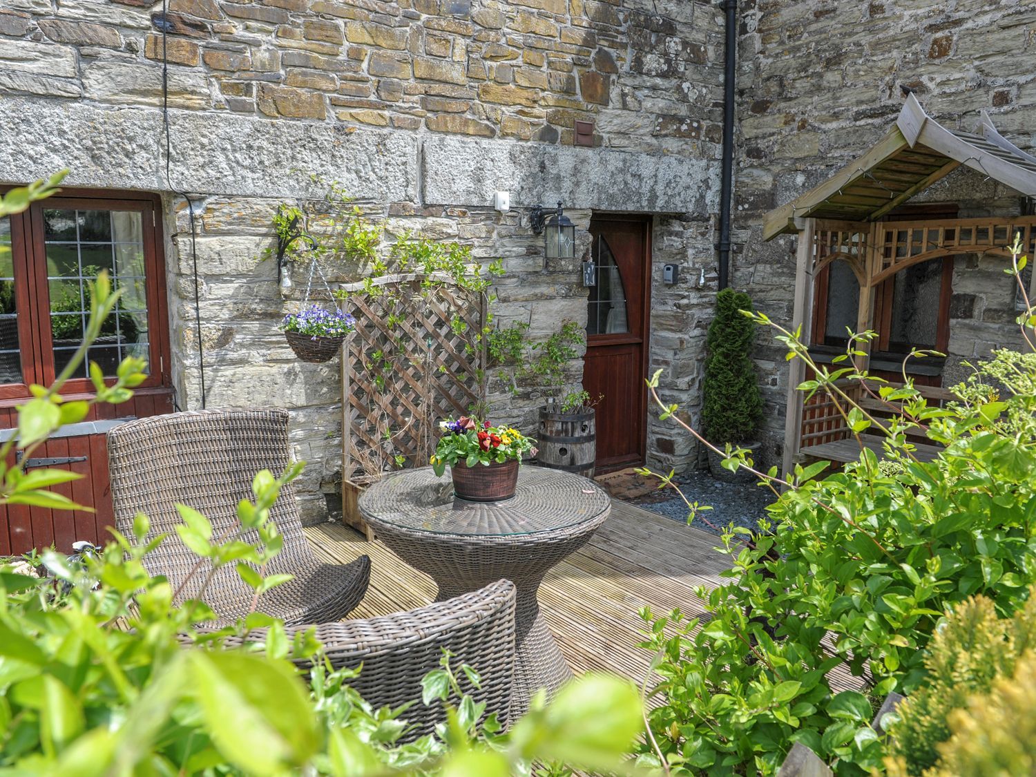 An outdoor patio with wicker chairs a table with flowers and a wooden bench against a stone wall at Nightingale Cottage in Lostwithiel