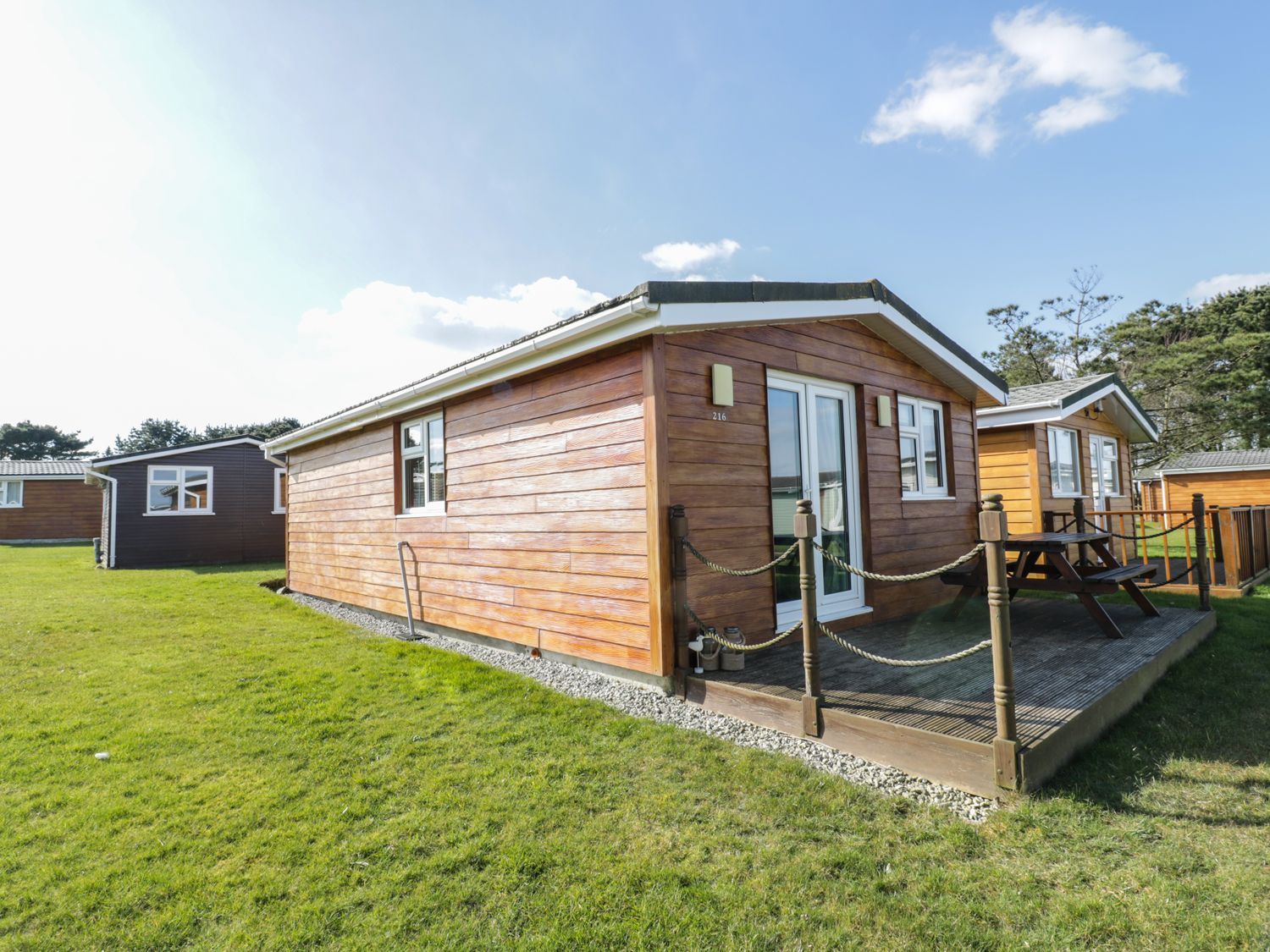 A wooden cabin with a small deck and picnic table on grass at Chalet 216- The Beach Retreat Atlantic Bays Holiday Park near St Merryn