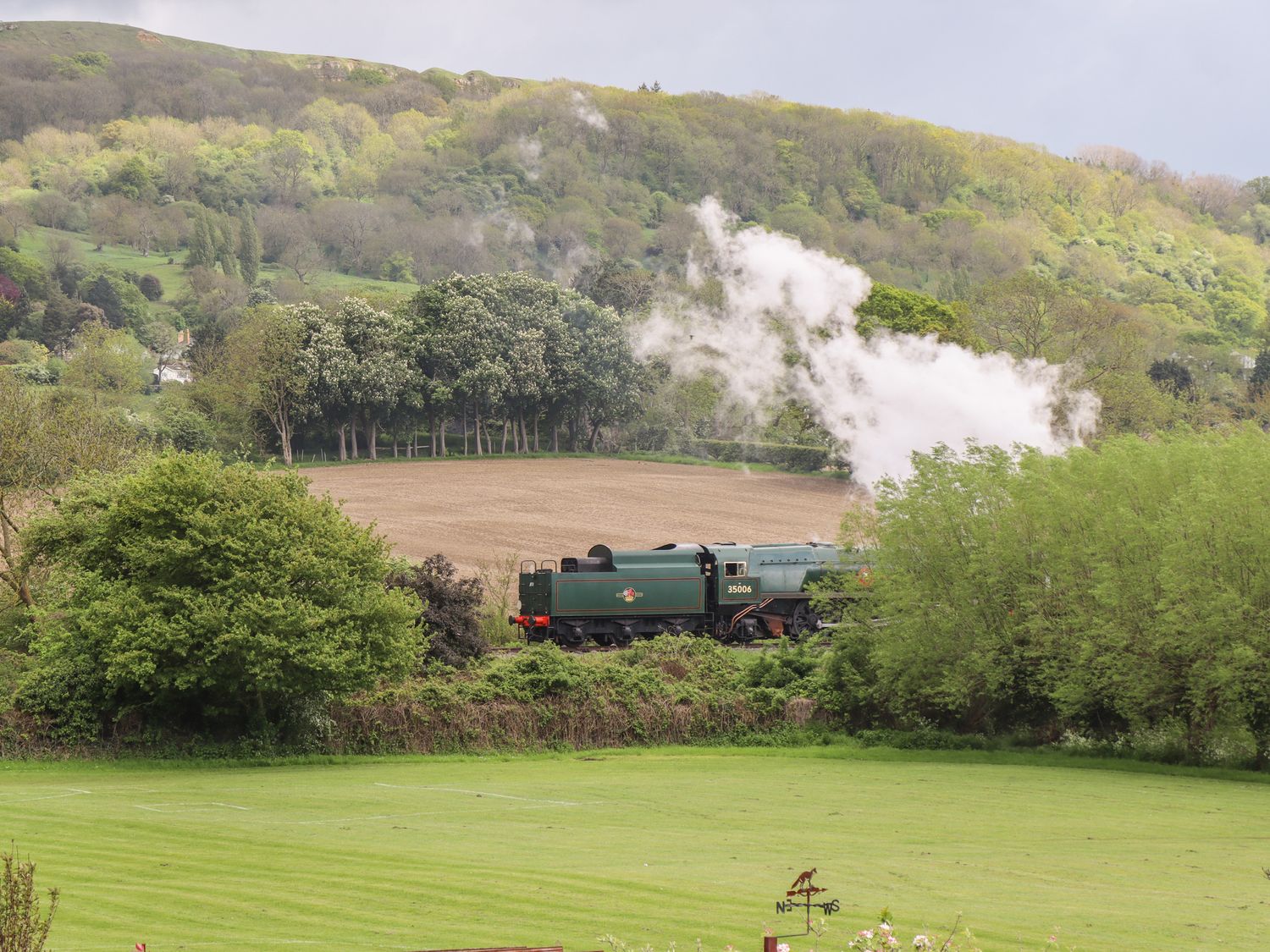 A green steam train emitting smoke traveling through a countryside landscape with fields trees and hills