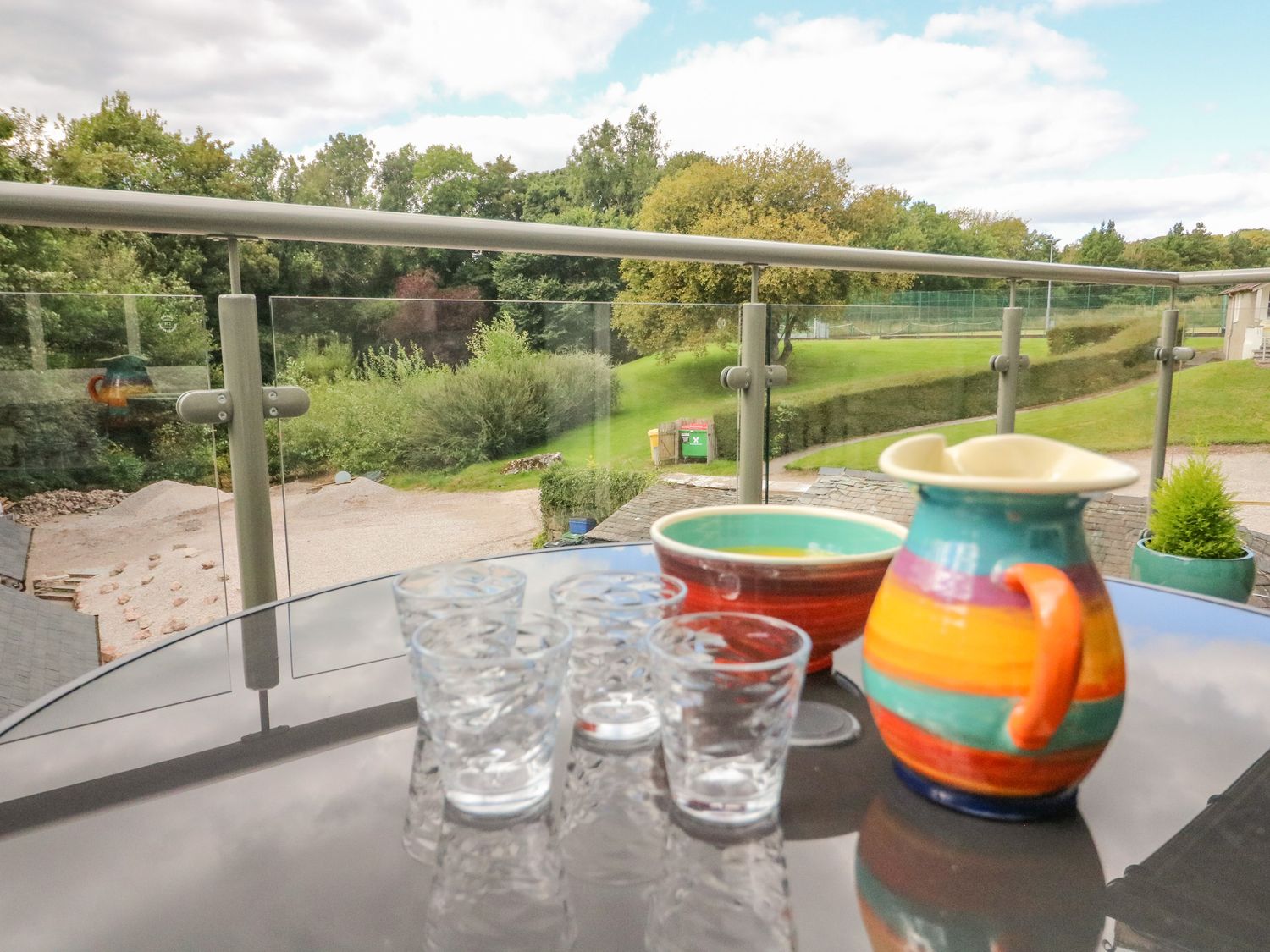 A balcony with a glass table holding four clear glasses and a colorful jug and bowl overlooking a grassy area with trees at Bronte House in Kirkby Lonsdale