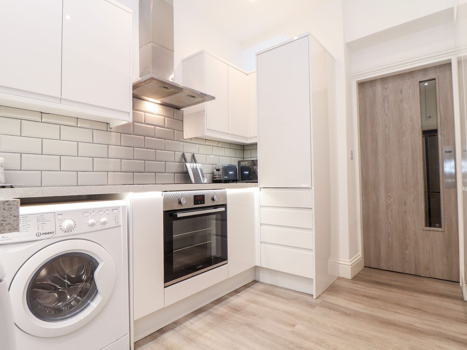 A kitchen with white cabinets washing machine oven toaster and wooden door at Teylu Camborne