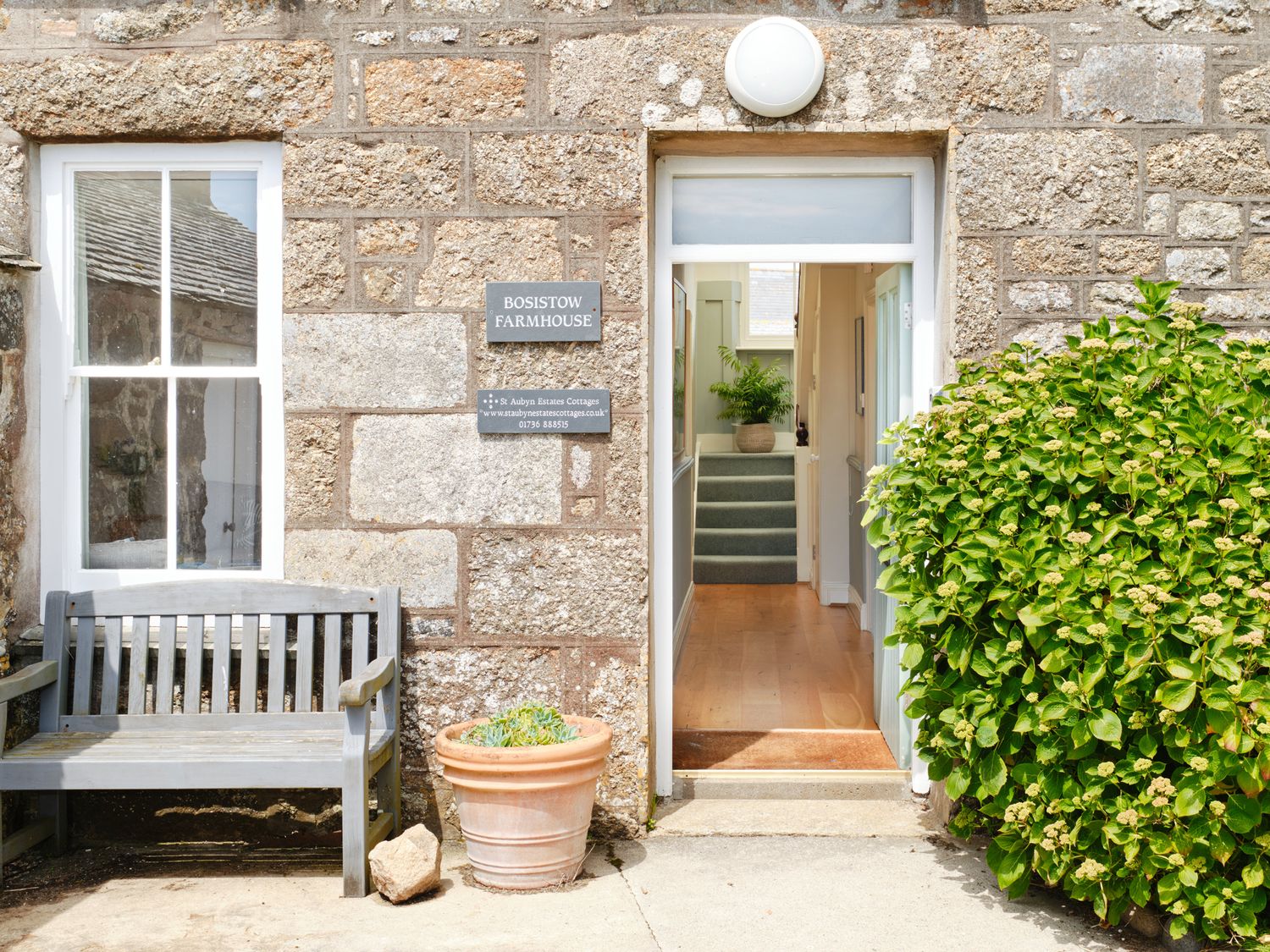 The exterior entrance of Bosistow Farmhouse with a wooden bench, a potted plant, a green bush, and an open door showing a hallway with stairs
