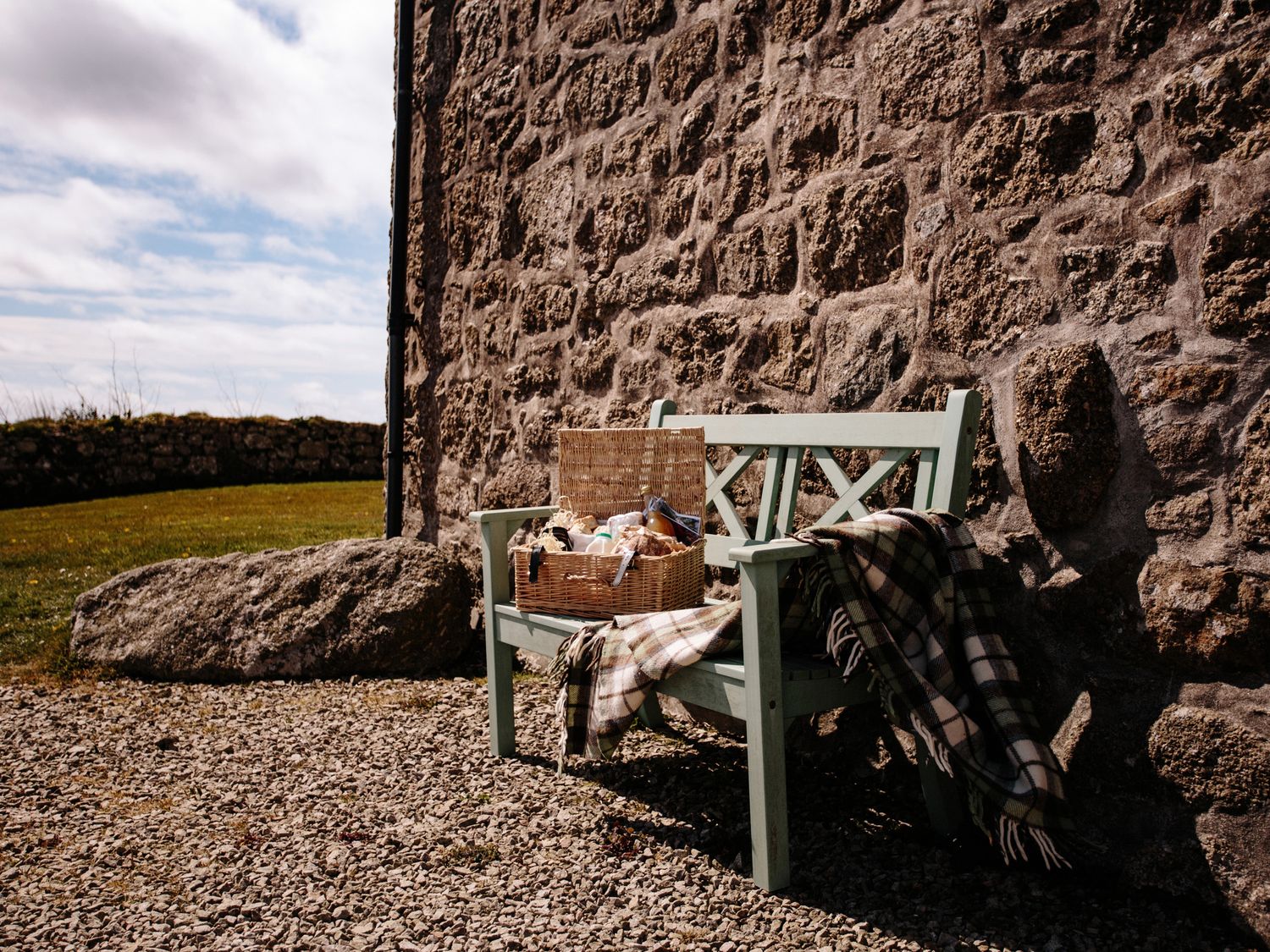 A green wooden bench with a picnic basket and a plaid blanket beside a stone wall outdoors