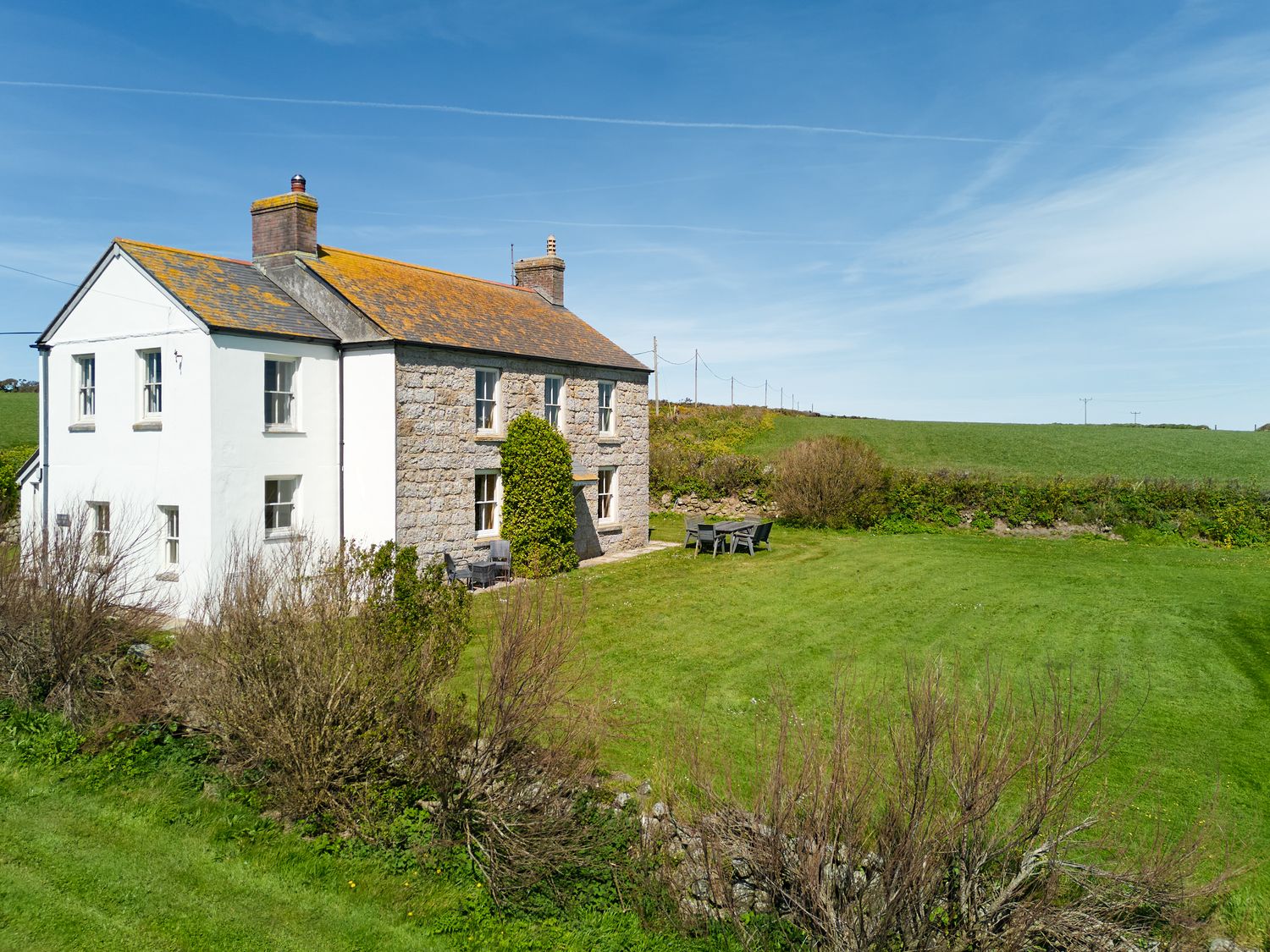 A stone and white painted house with two chimneys and garden furniture on a lawn at Higher Roskestal in St Levan near Porthgwarra