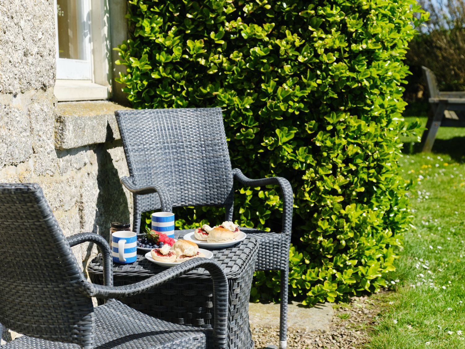 An outdoor seating area with two wicker chairs and a table with plates of scones and mugs near a stone wall and hedge at Higher Roskestal in St Levan near Porthgwarra