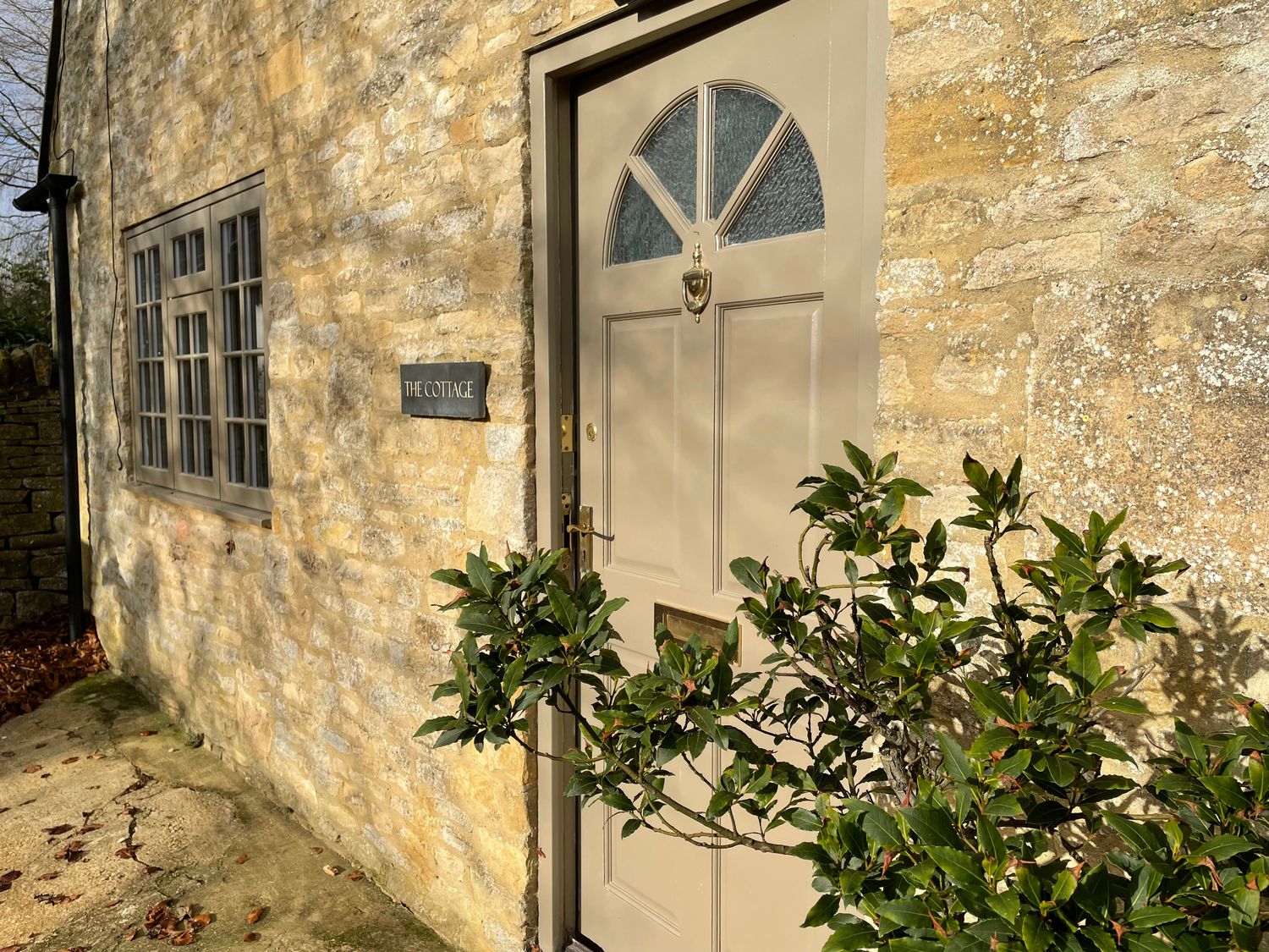 An exterior view of a stone cottage with a beige wooden door a window and a small plant nearby at Omas Cottage Moreton-In-Marsh