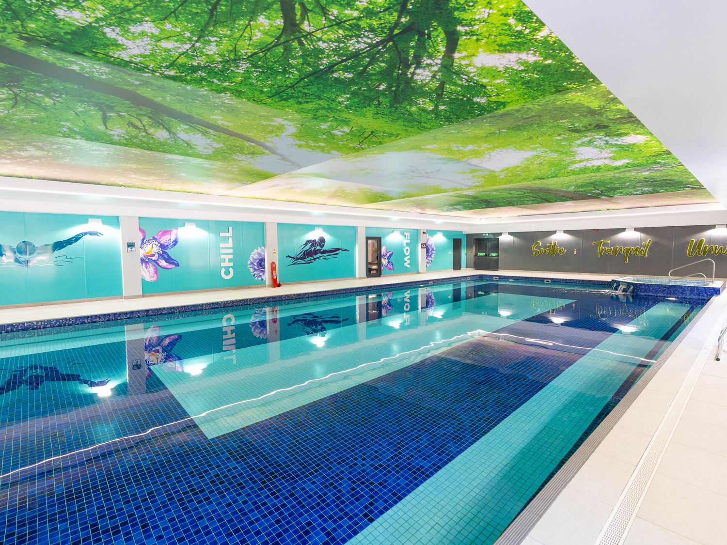 An indoor swimming pool with blue tiles and wall murals under a ceiling with tree images at Bracken Cottage in St Tudy