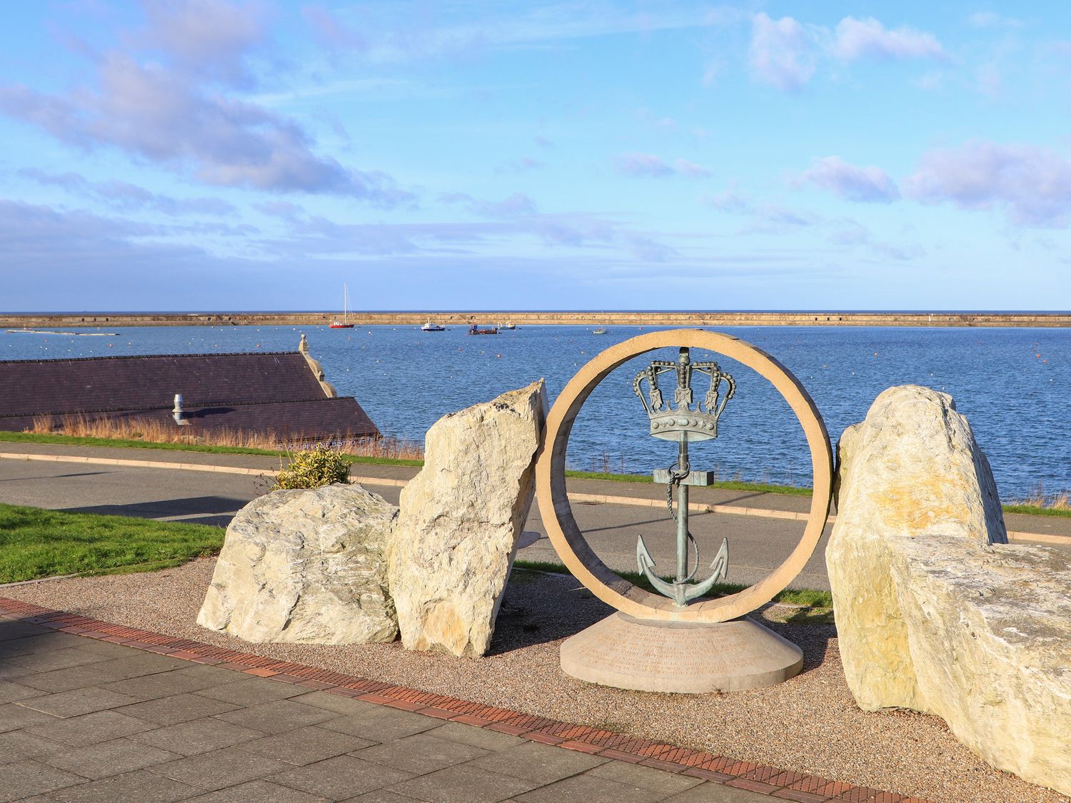 A circular monument with an anchor and crown framed by large stones near a waterfront with boats at Swn Y Nant in Holyhead
