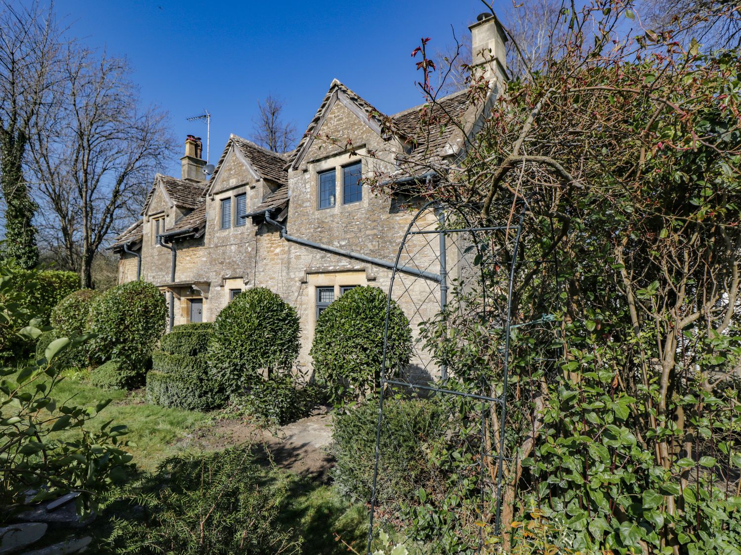 A stone cottage with multiple gables and windows surrounded by trimmed bushes and trees at Rose Cottage in Corsham