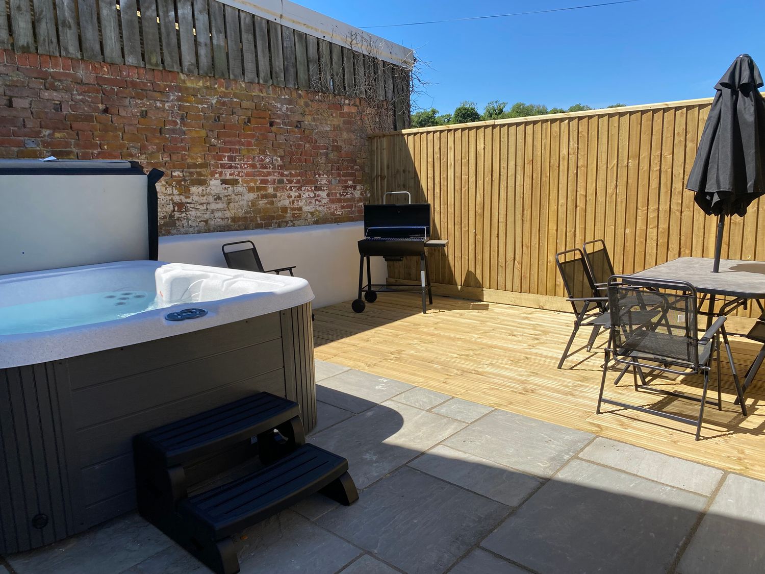 An outdoor patio area with a hot tub, a barbecue grill, a table with chairs and an umbrella at The Mill in Thornicombe near Blandford Saint Mary