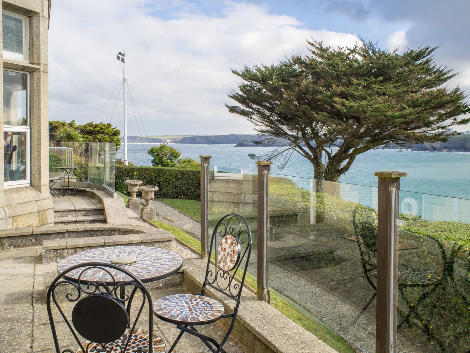 A patio with mosaic table and chairs overlooking a tree hedge and sea at King Edward's View in Newquay