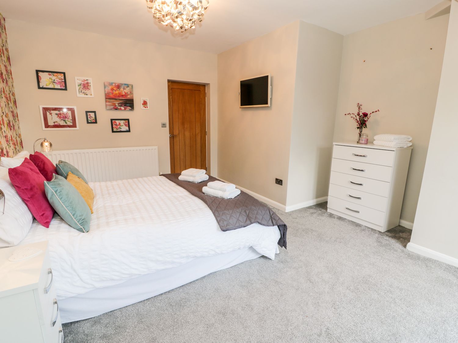 A bedroom with a bed pillows a wooden door a dresser with flowers and a wall-mounted television at The Chalet in Benllech