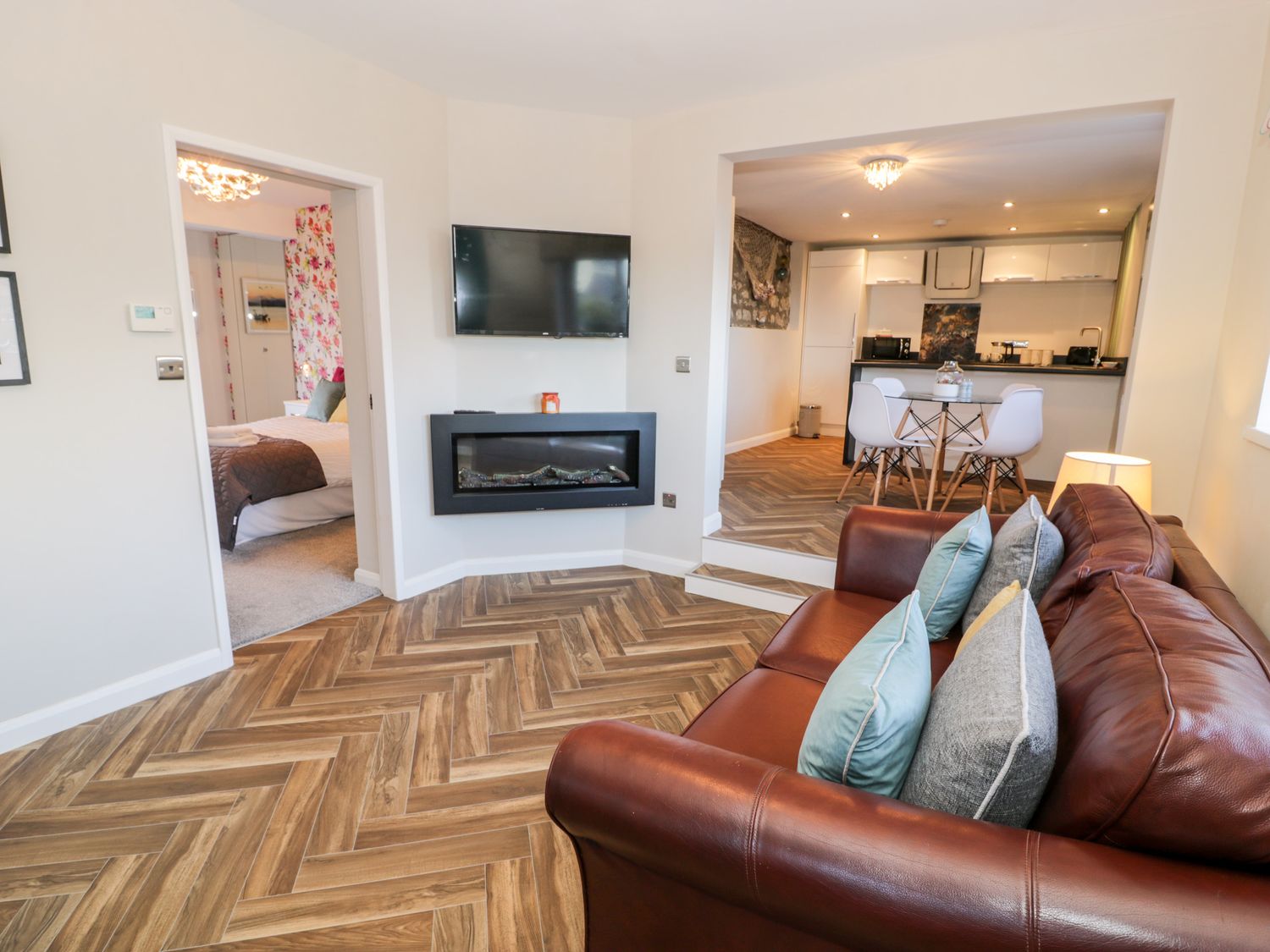 A living room with a brown leather sofa and pillows a wall mounted TV a fireplace an open doorway to a bedroom and an open kitchen and dining area at The Chalet in Benllech