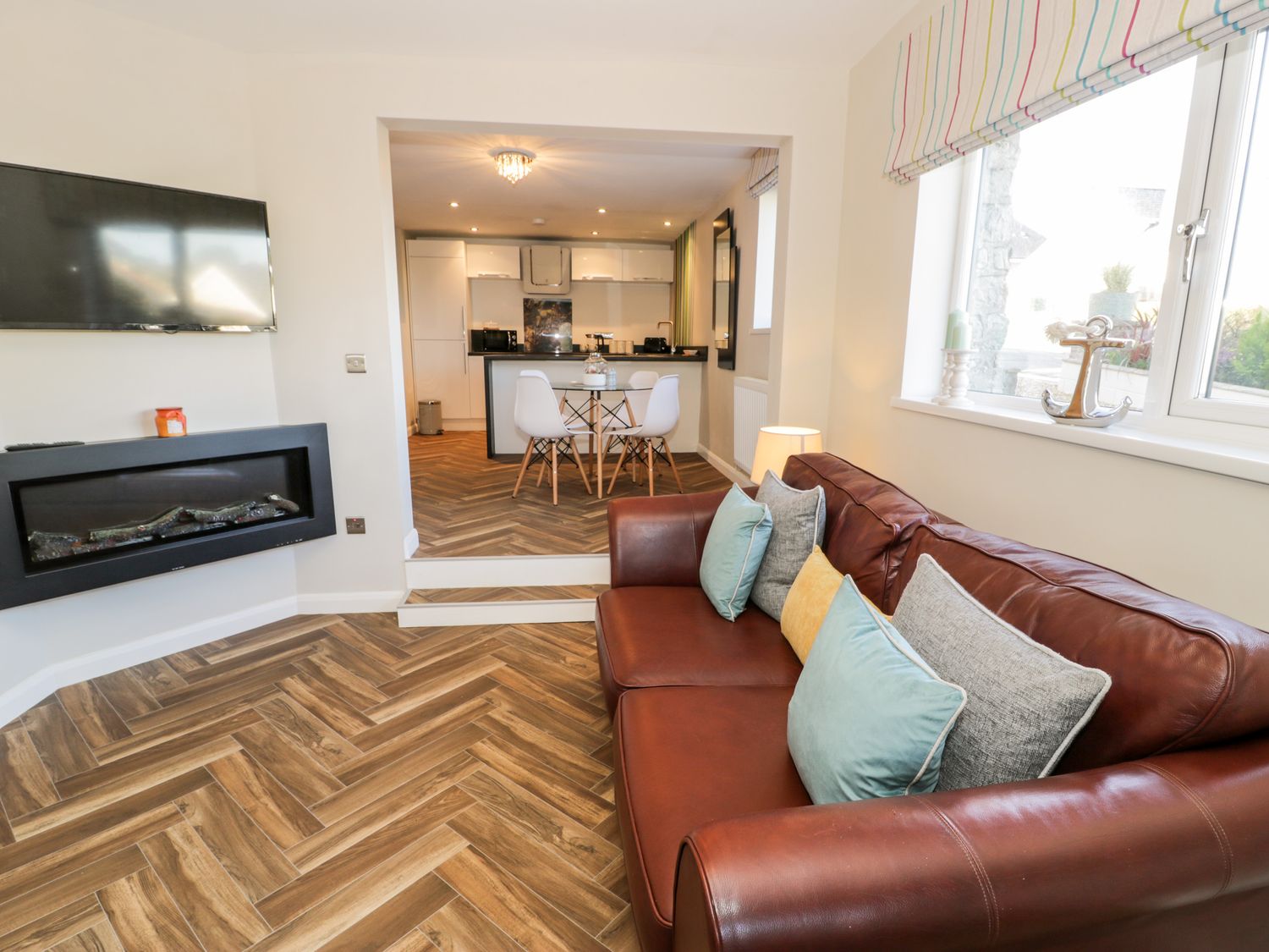 A living room with a brown leather sofa and cushions a wall-mounted tv and a fireplace with a view to a dining area and kitchen at The Chalet in Benllech