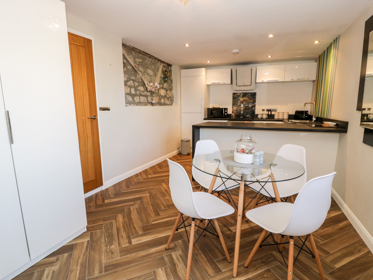 A kitchen with a glass dining table and four white chairs a wooden door and white cabinets at The Chalet in Benllech