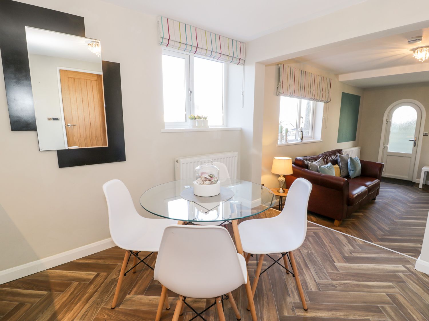 A dining area with a round glass table and white chairs next to a living room with a brown leather sofa and lamp at The Chalet in Benllech