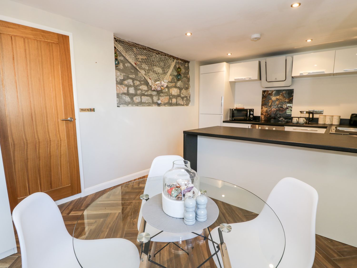 A kitchen with white cabinets and appliances next to a glass dining table with white chairs at The Chalet in Benllech