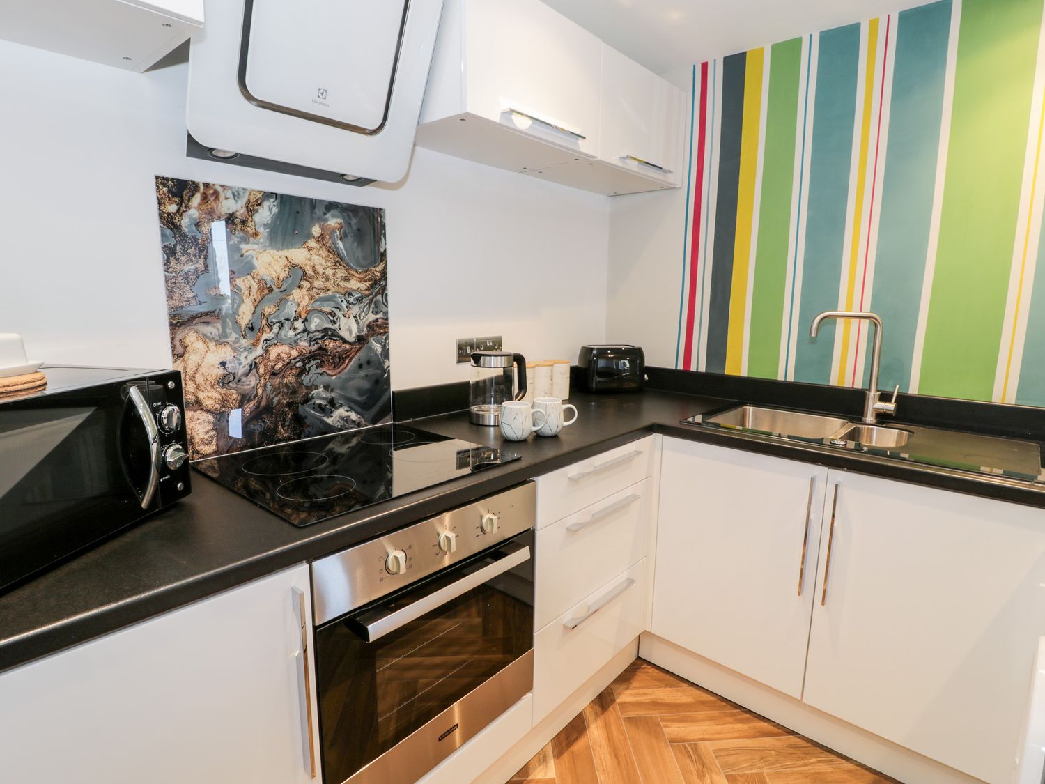 A kitchen with white cabinets a stove microwave kettle two mugs and a sink with a striped wall at The Chalet in Benllech