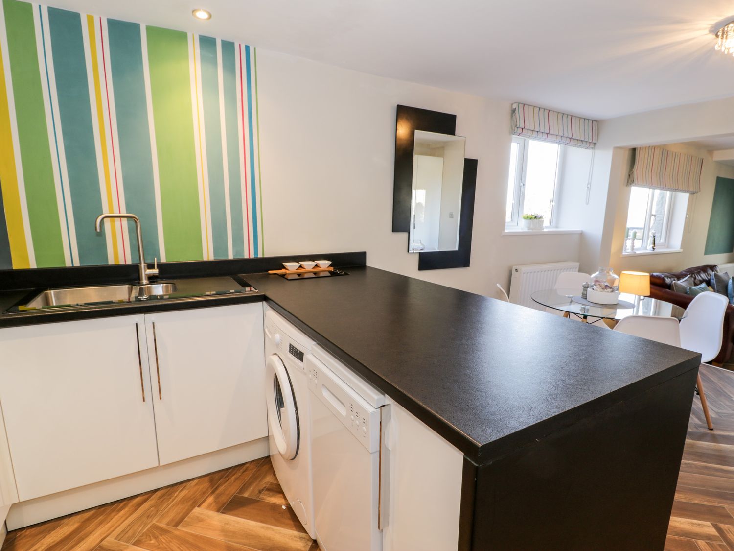 A kitchen with a black countertop white cabinets washing machine and dishwasher a dining area with white chairs and table at The Chalet in Benllech