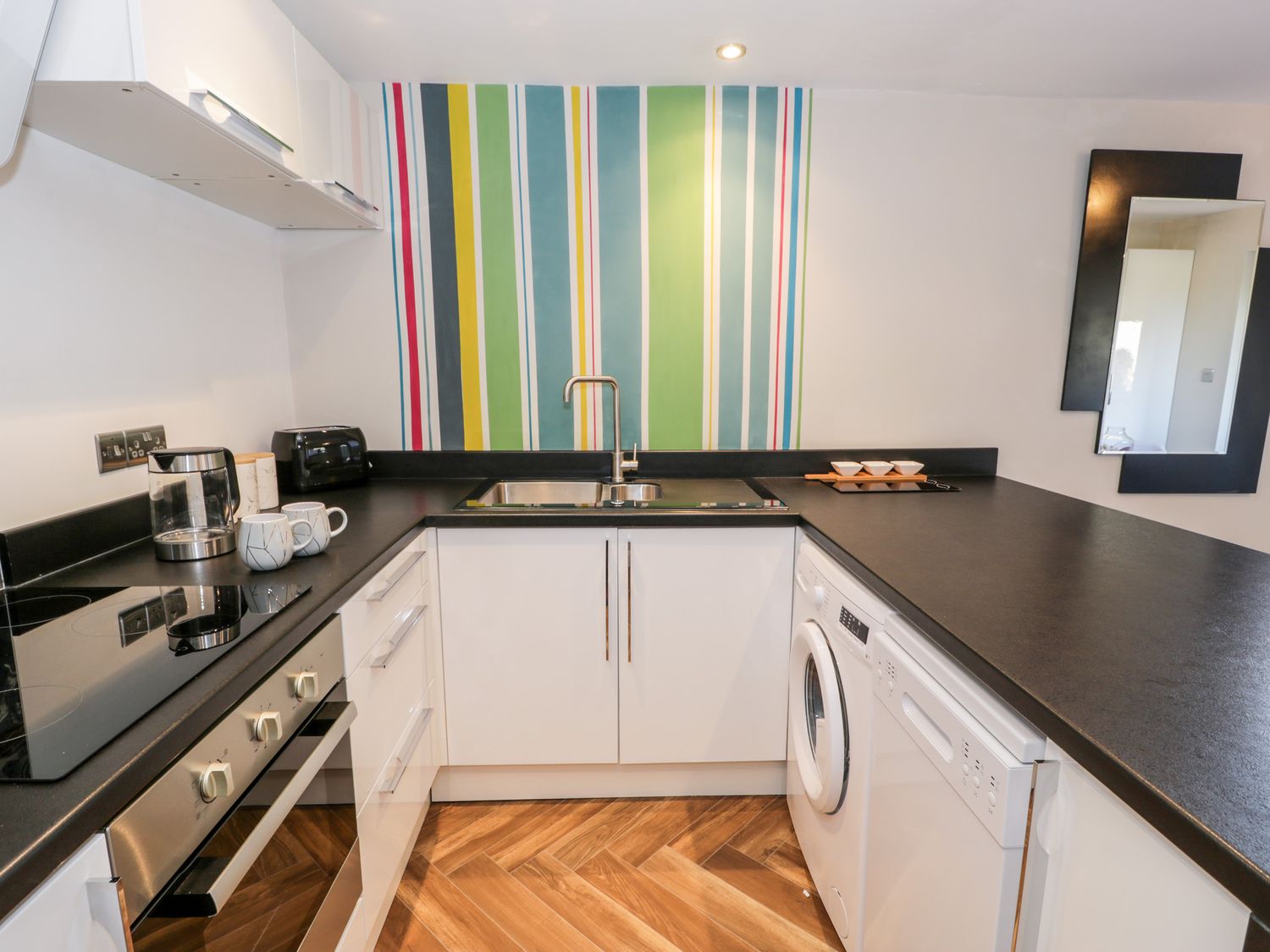 A kitchen with white cabinets black countertops a stove oven sink washing machine and colorful striped wall at The Chalet in Benllech