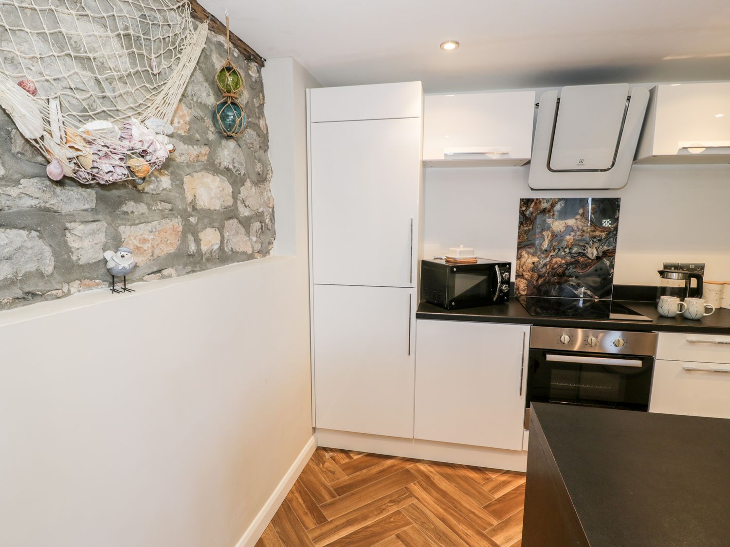A kitchen with white cabinets black countertops a microwave oven and a stone wall with shells at The Chalet in Benllech