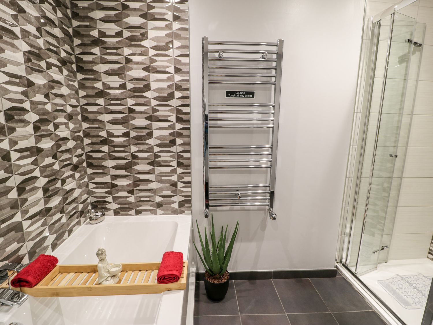A bathroom with a bathtub with a wooden tray and red towels a chrome towel rail a potted plant and a glass shower enclosure at The Chalet in Benllech