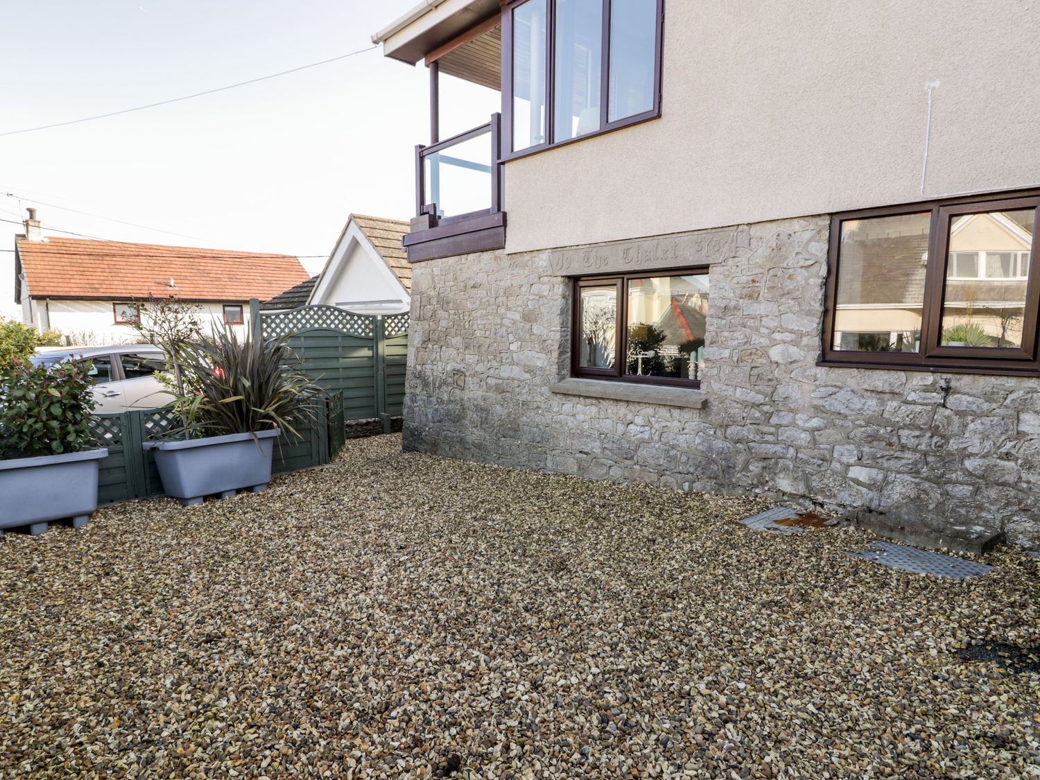 A gravel yard with potted plants and green fence panels beside a stone and stucco house with windows at The Chalet in Benllech