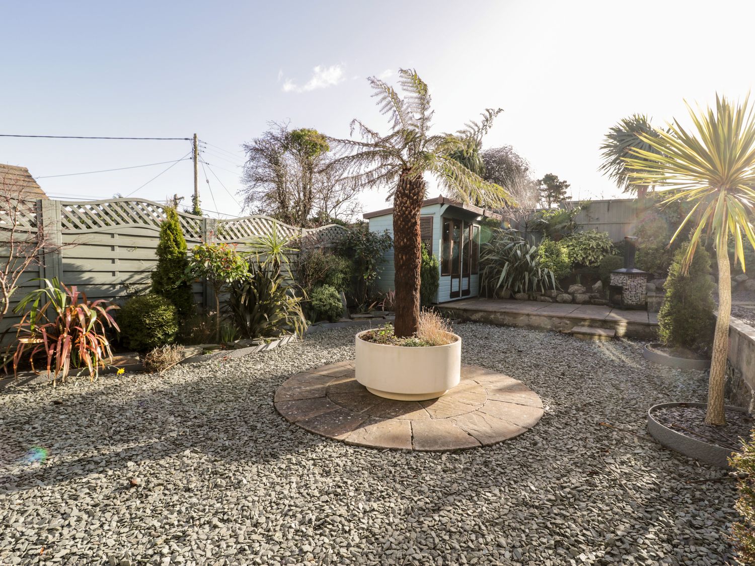 A garden with gravel ground a potted tree in the center plants along a fence and a garden shed at The Chalet in Benllech