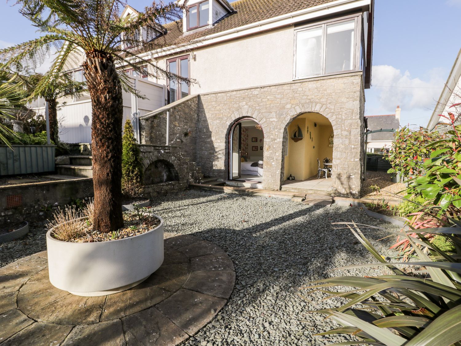 An outside view of a stone house with arched doorways and a garden area with gravel and a tree in a planter at The Chalet in Benllech