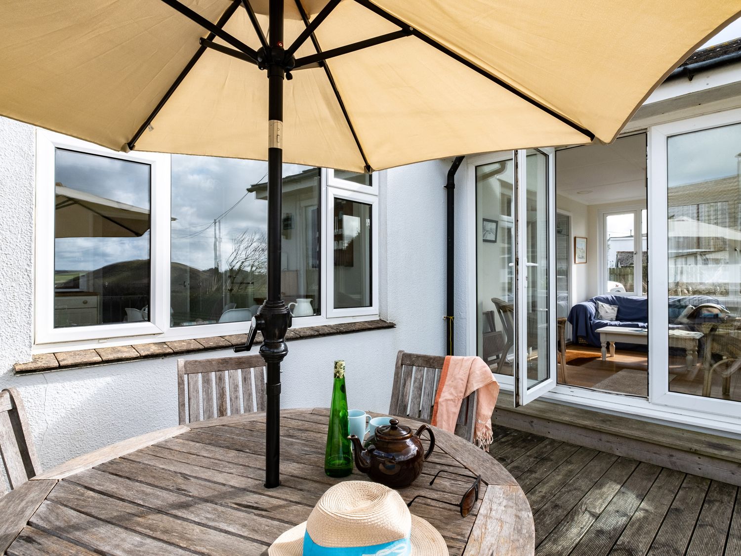 An outdoor patio with a round wooden table and chairs under a beige umbrella at Croft Elms in Trebetherick near Polzeath