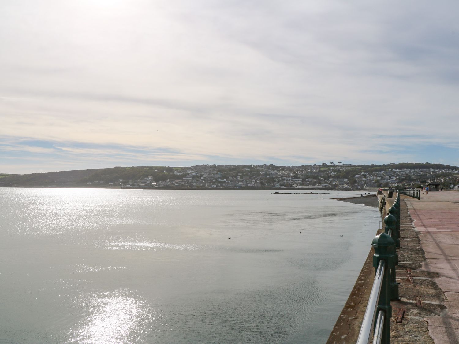 A waterfront promenade with railings along calm water and houses on a hillside in the distance at Rose Bank Cottage in St Just