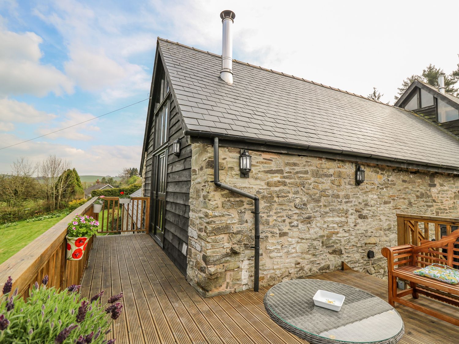 An outdoor wooden deck attached to a stone and wood house with potted plants a round glass table and wooden bench with cushions at Goblaen Barn in Rhosgoch near Clyro