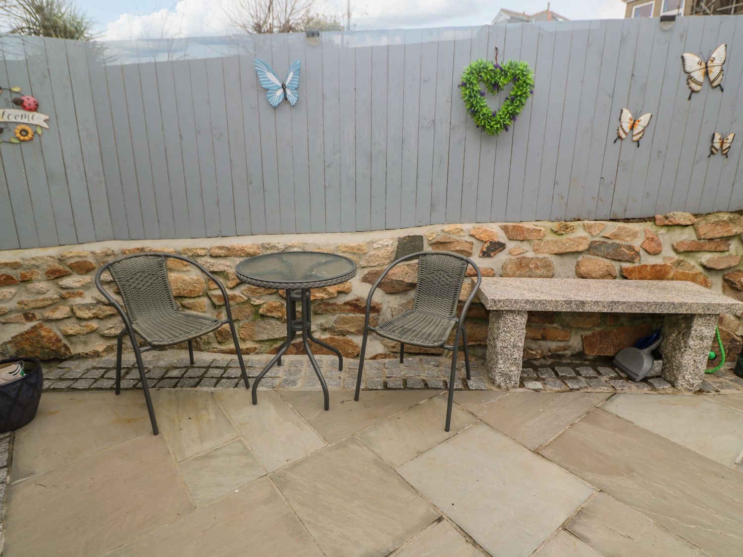 An outdoor seating area with two chairs a round table a stone bench and a gray wooden fence with butterfly and heart decorations at Pippas place in St Just