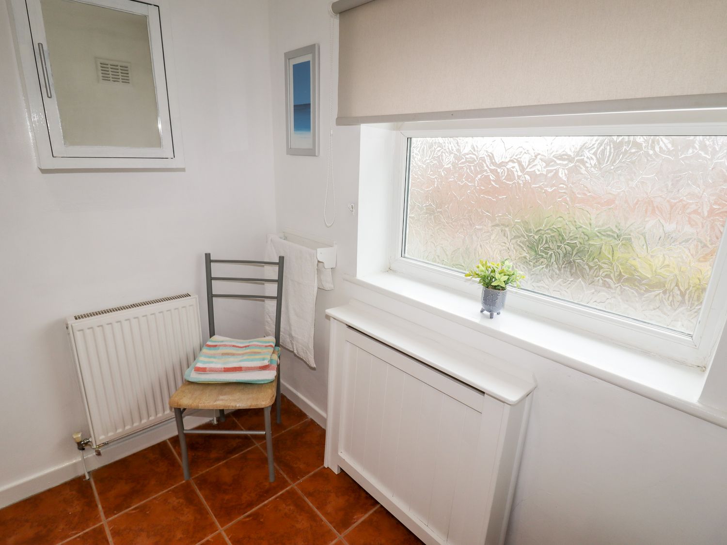 A small room with a chair holding folded towels a radiator a towel rack a frosted window and a small plant on the windowsill at The Hawthorns in Christleton
