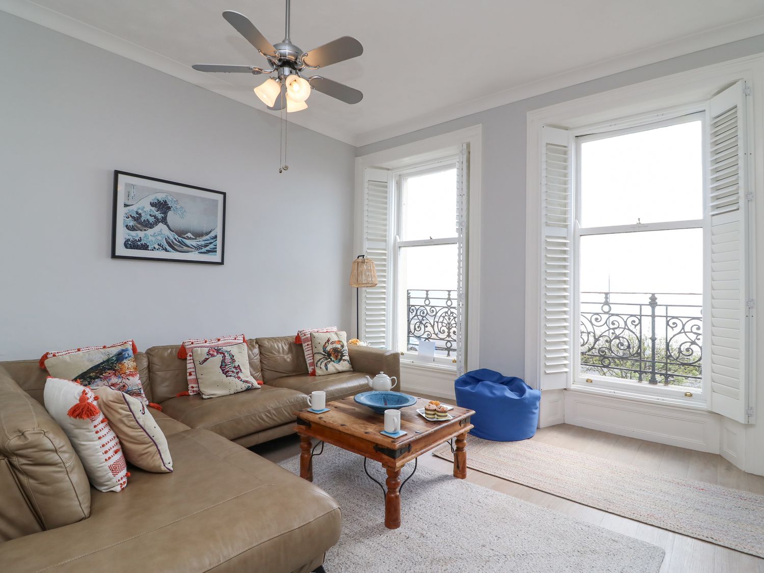 A living room with a brown sectional sofa and decorative pillows a wooden coffee table with cups and a teapot two large windows with white shutters and a blue bean bag at Sunrise Apartment in Weymouth