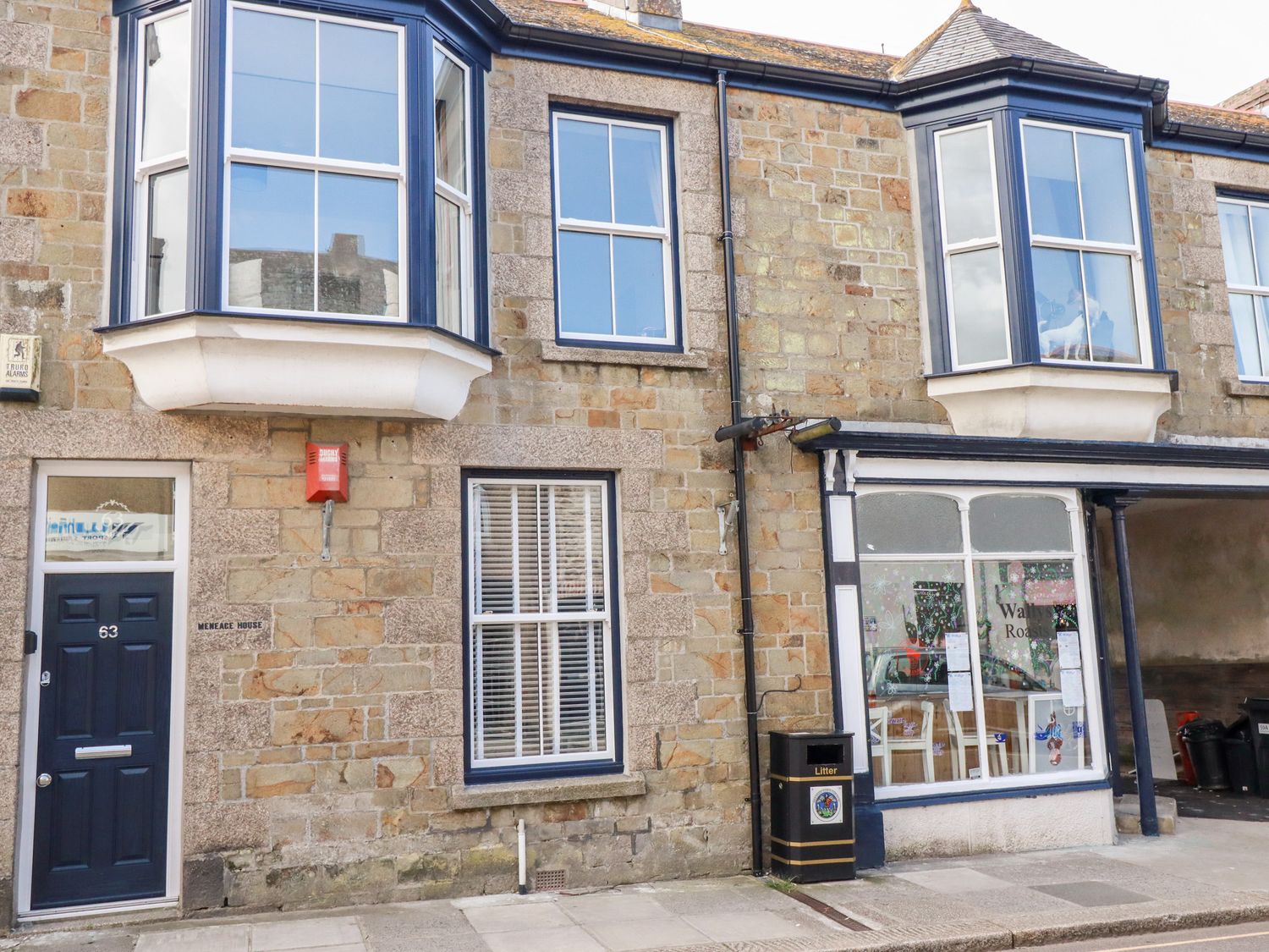 Stone building with blue framed windows a blue door labeled 63 and a shop window with litter bin outside at Meneage House Helston