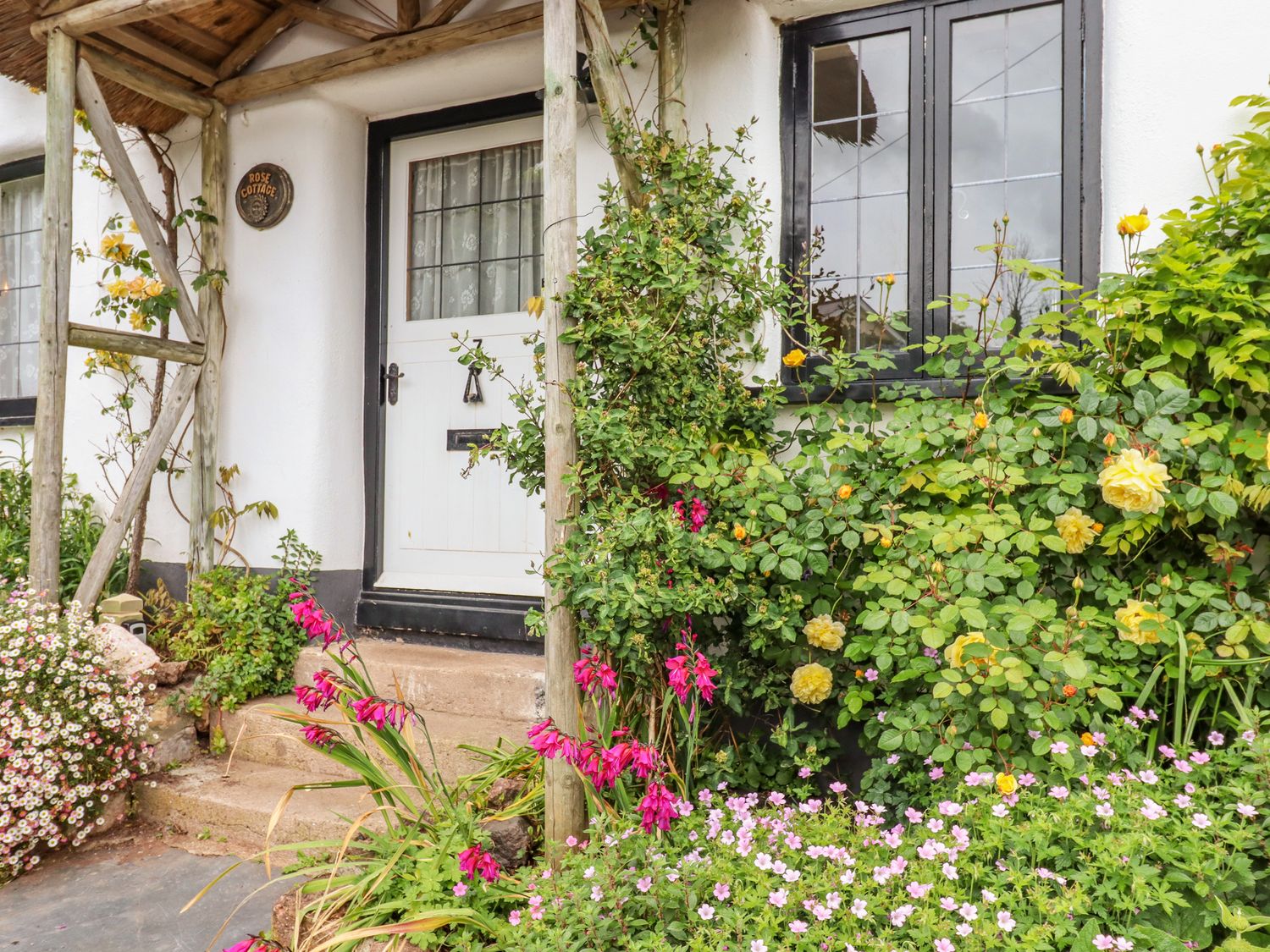 The entrance to a cottage with a white door black framed windows and flowers in the garden at Rose Cottage in Holcombe in Teignmouth