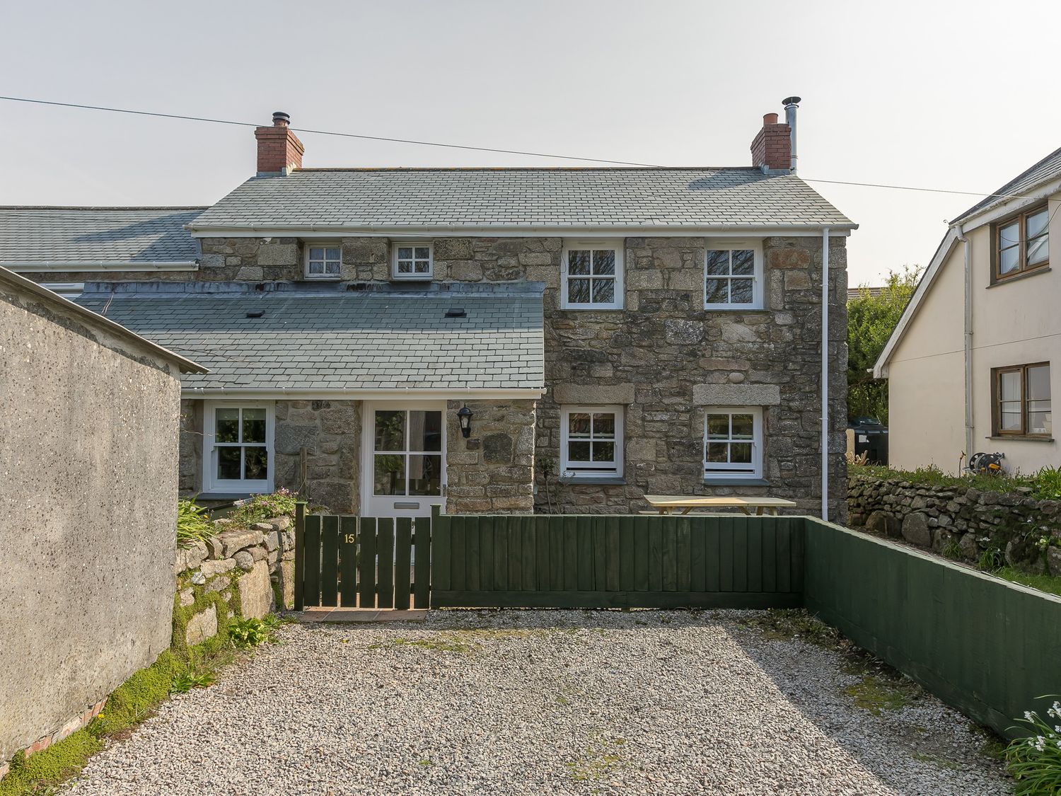 A stone cottage with white windows and two chimneys behind a green wooden fence and gravel area at 15 Boscaswell Downs in Pendeen