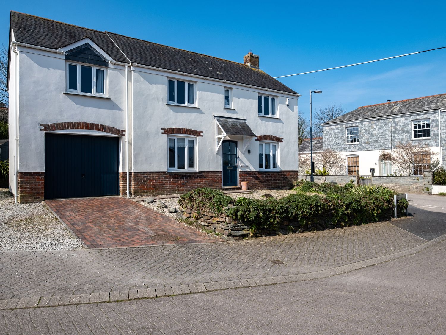 A detached house with white walls and a brick base on a corner with a driveway and shrubs at Marbeachow in Tredrizzick near Rock