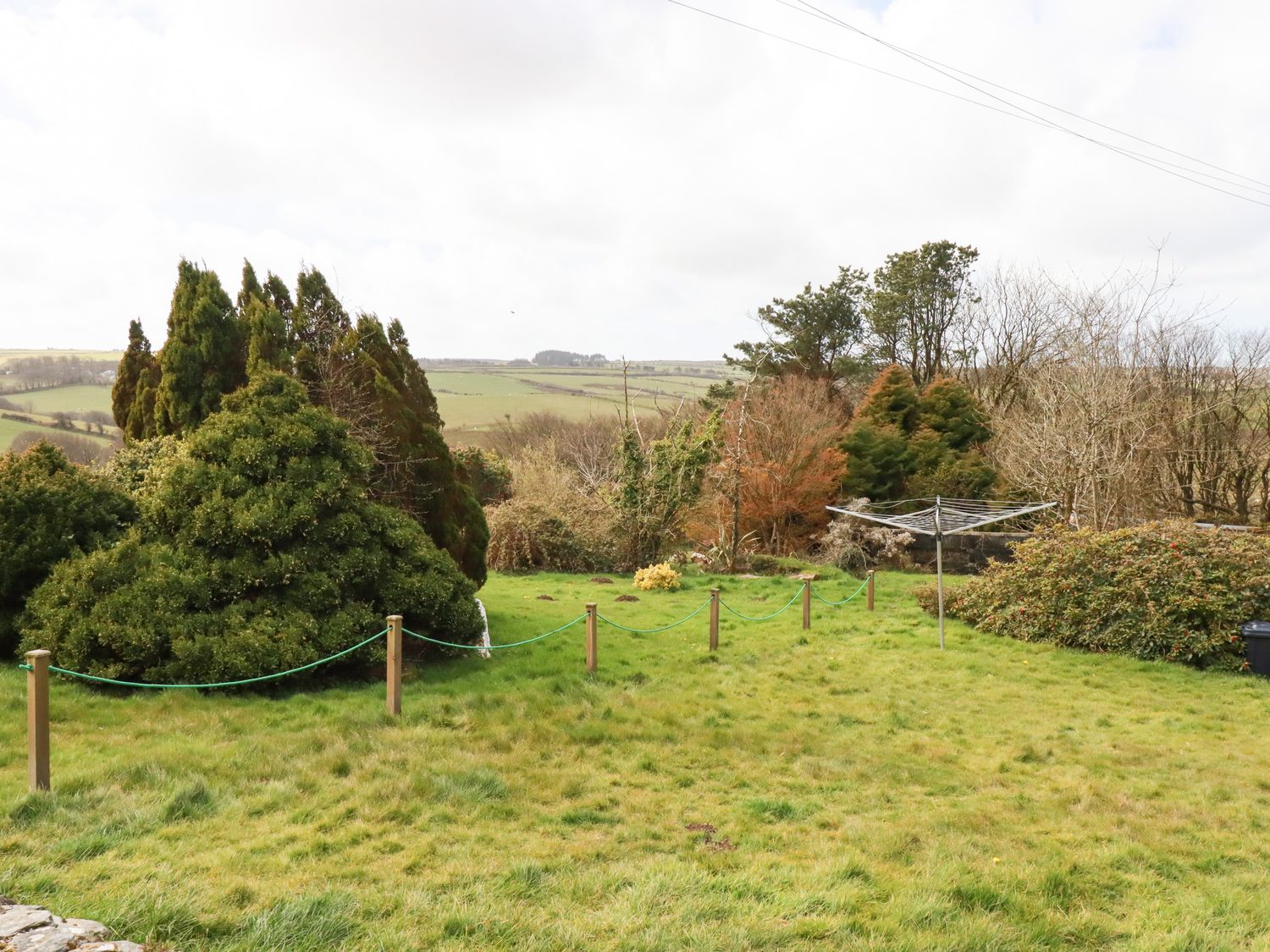 A grassy garden area with wooden posts connected by green rope bushes and trees in the background at Brightwell Barn Treglasta near Hallworthy