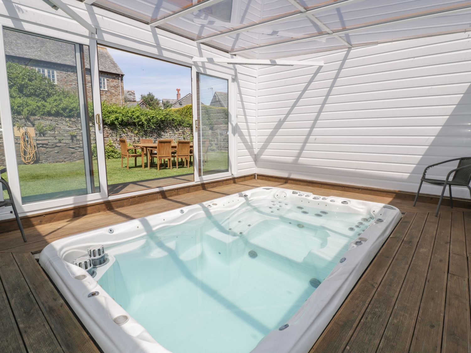 A covered patio area with an indoor hot tub and glass sliding doors opening to a garden with a wooden table and chairs at Owls Roost in Gorran Haven