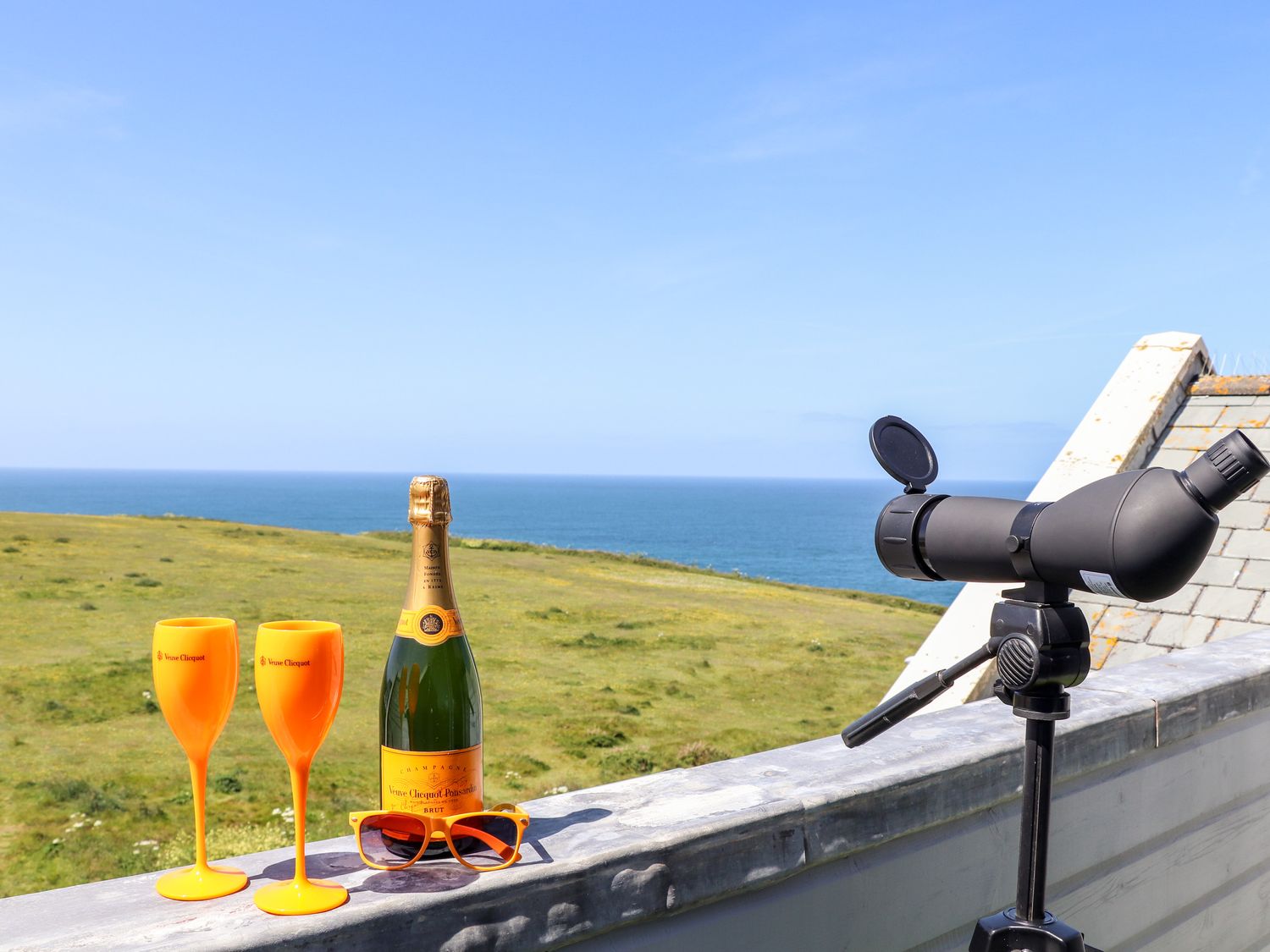 A balcony with a bottle of champagne two orange glasses orange sunglasses and a telescope overlooking the sea and grassy field at Penthouse 23 in Newquay