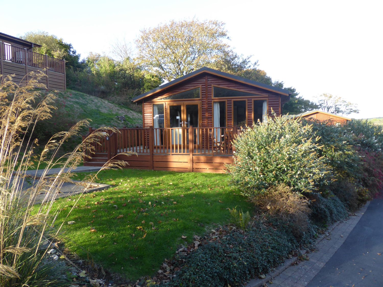 A wooden cabin with a fenced porch surrounded by grass and bushes at 23 Bossiney Bay in Tintagel