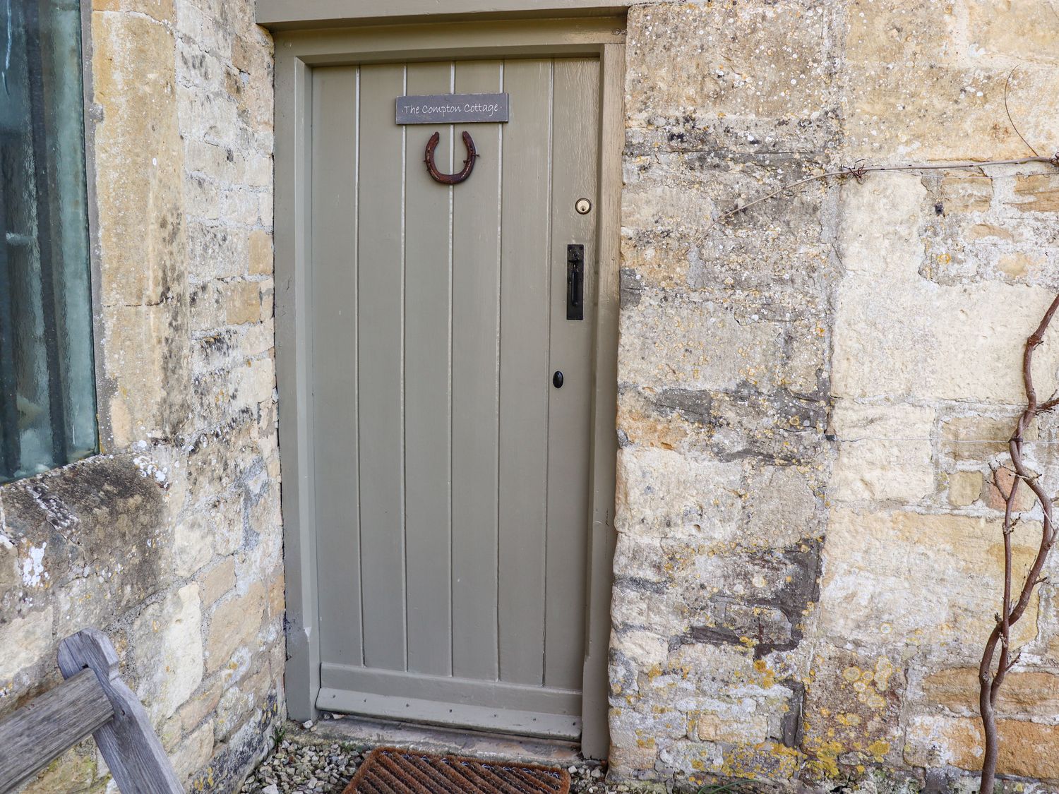 A grey wooden door with a horseshoe and sign reading The Compton Cottage set in a stone wall at The Compton Cottage in Northleach