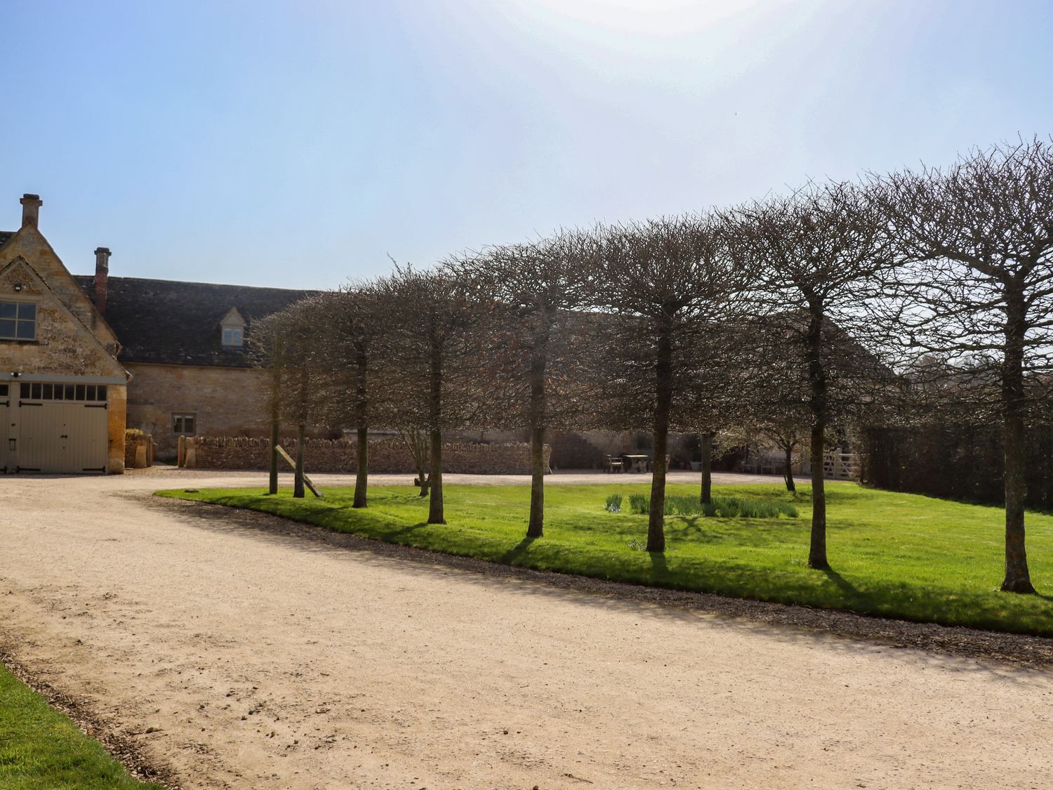 A gravel driveway with trimmed trees and a stone house in the background at The Compton Cottage in Northleach