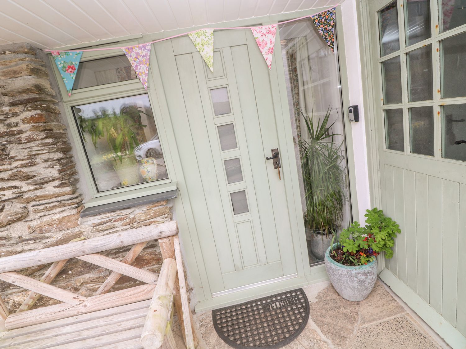 A front door with a welcome mat and plants next to a wooden bench at Wheel Cottage in Pelynt