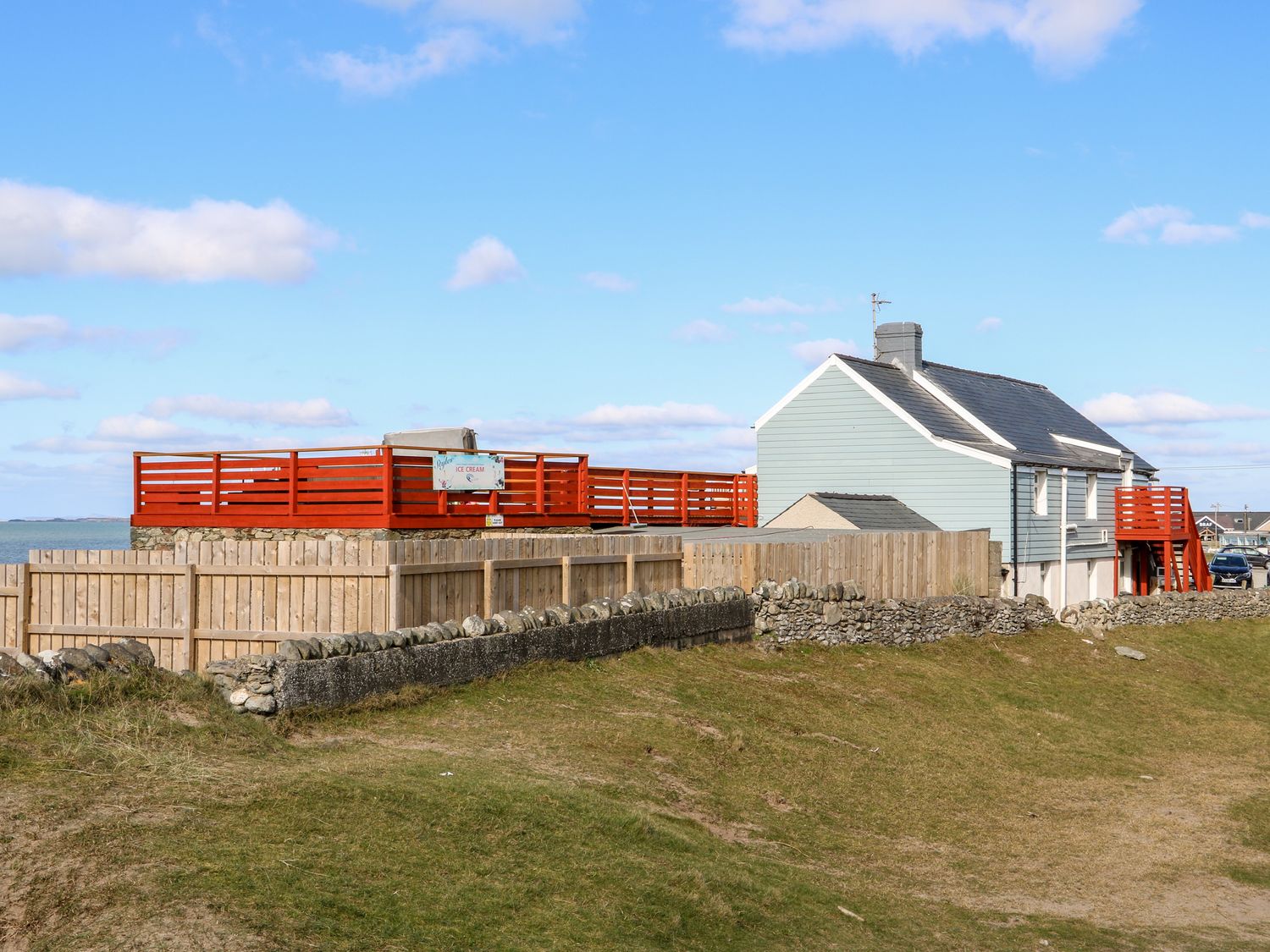 A blue house with a fenced yard and red wooden deck near the sea at Swn Y Mor in Dinas Dinlle