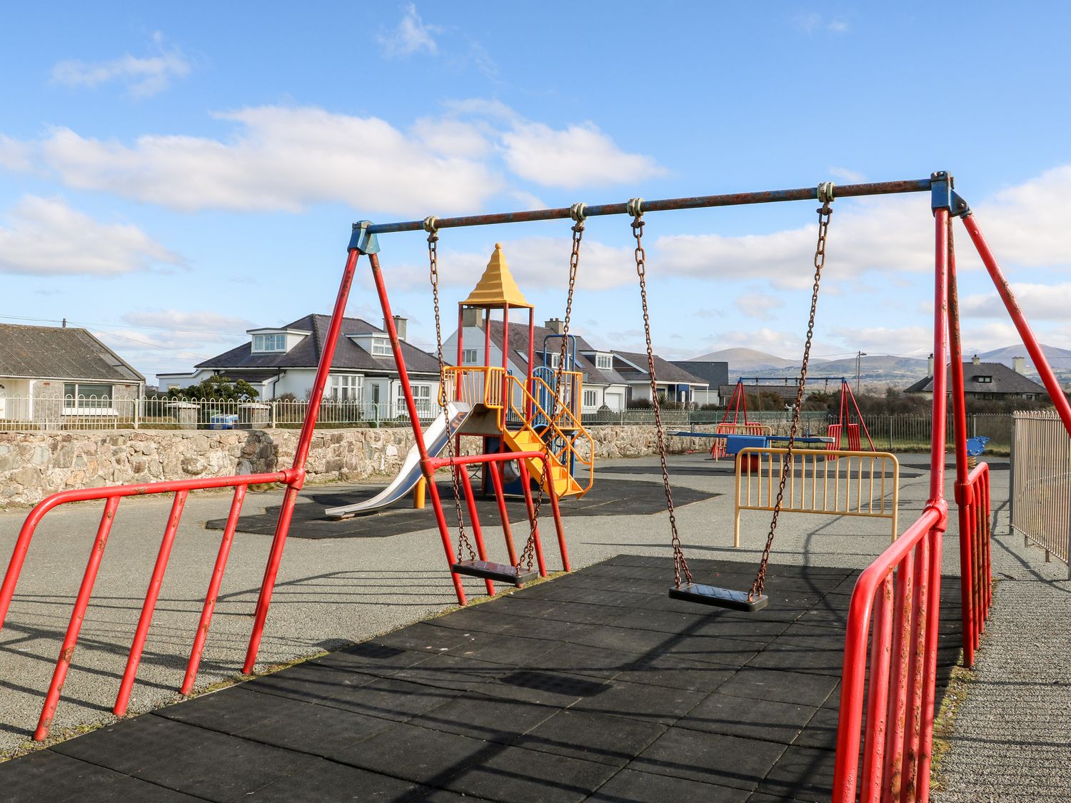 An outdoor playground with red swings and a slide near houses at Swn Y Mor in Dinas Dinlle