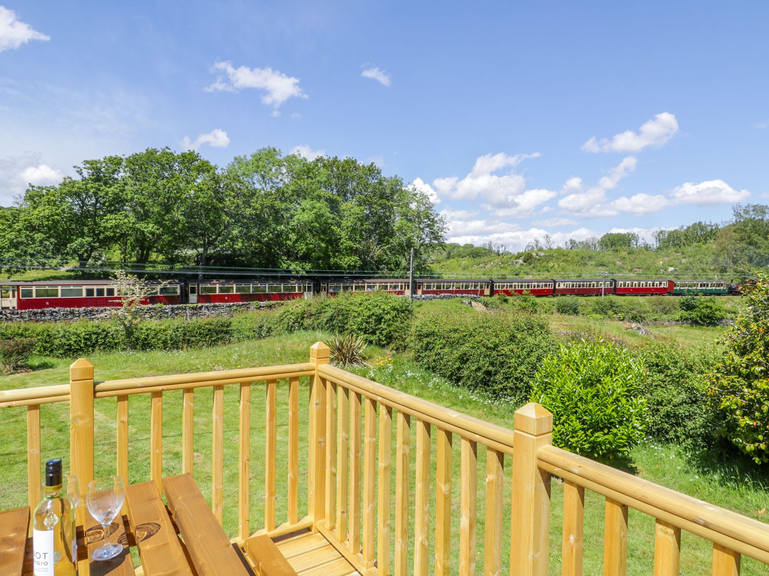 A wooden deck with a table holding a bottle and glasses overlooking a grassy area with bushes and a train passing in the background at Ceris in Penrhyndeudraeth