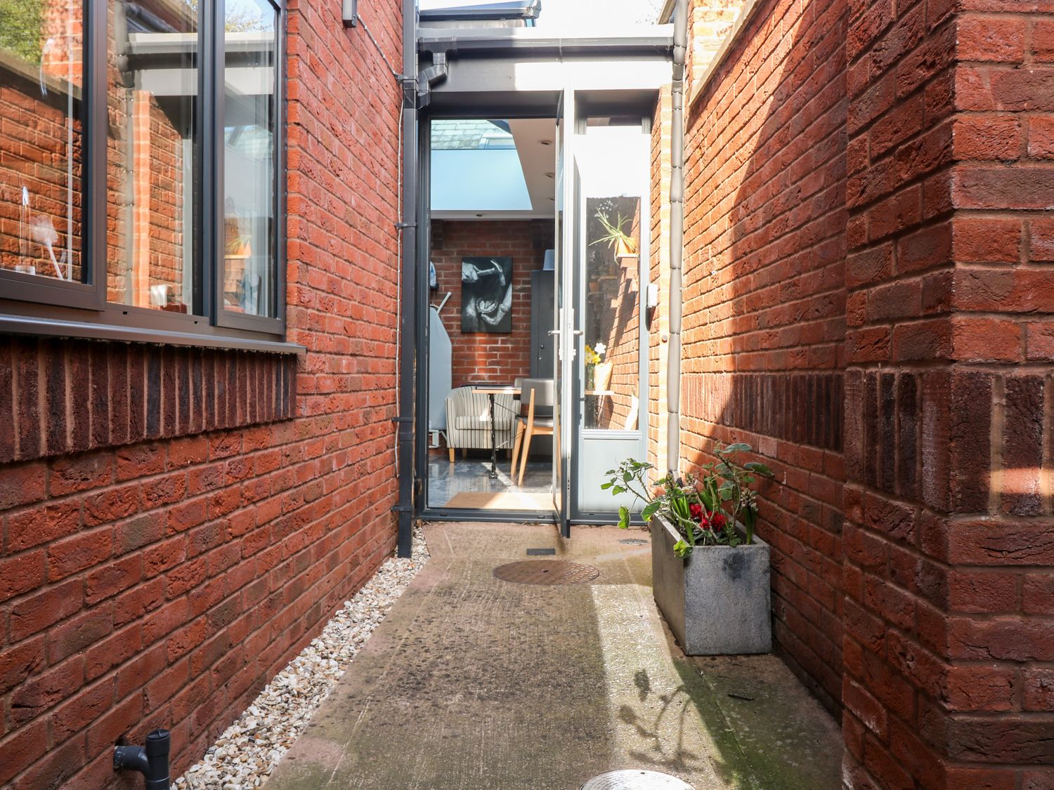 A narrow outdoor corridor with red brick walls on both sides a concrete planter with plants and an open glass door leading to a room with chairs at Farriers Retreat Budleigh Salterton